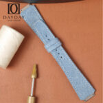Blue denim watch strap for RM 67 01 laid diagonally with white thread spool on a tan leather background