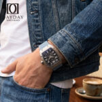 Man in denim jacket wearing Richard Mille RM 67 01 with blue denim watch strap by Daydaywatchband, casual street lifestyle wrist shot with coffee in background