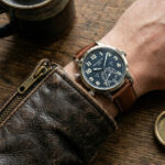 Patek Philippe Calatrava Pilot Travel Time 5524 on wrist with brown calfskin replacement strap, vintage leather wallet and compass on wooden table